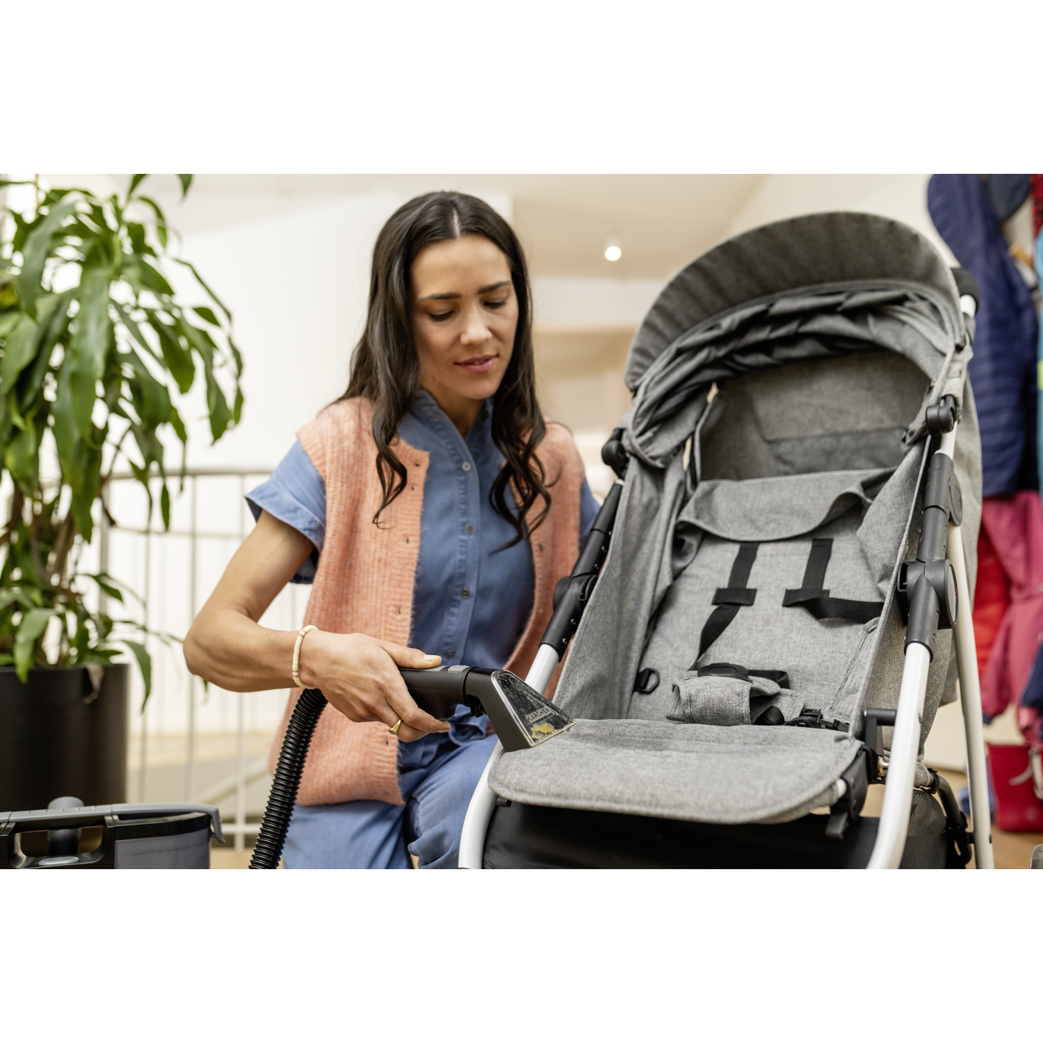 A woman cleans a gray stroller with a vacuum cleaner. She wears a blue dress and pink vest. A plant is in the background.