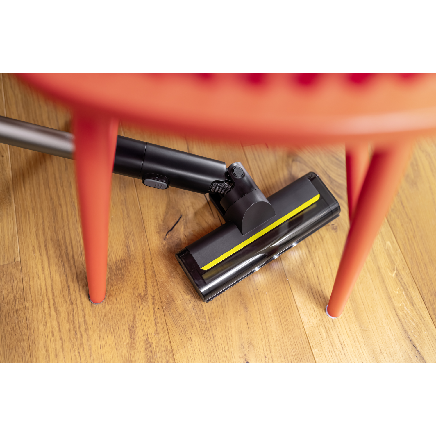 A black vacuum cleaner head with yellow details is under an orange chair. The floor is made of wood.