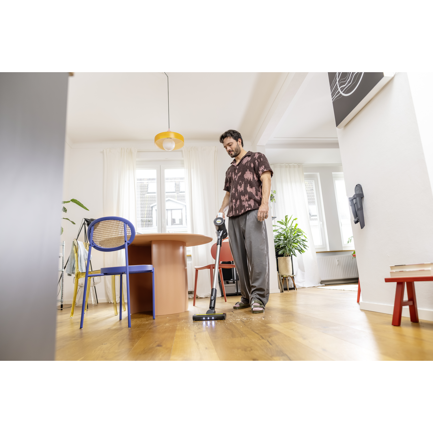 Man vacuums a wooden floor in a bright room. A round table and colorful chairs are in the background. There is a plant and a red stool.
