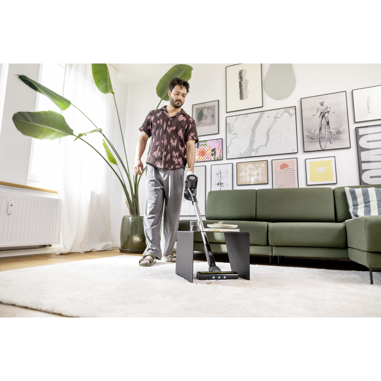 A man vacuums a white carpet in a living room. A green sofa, framed art, and a large plant are in the background.
