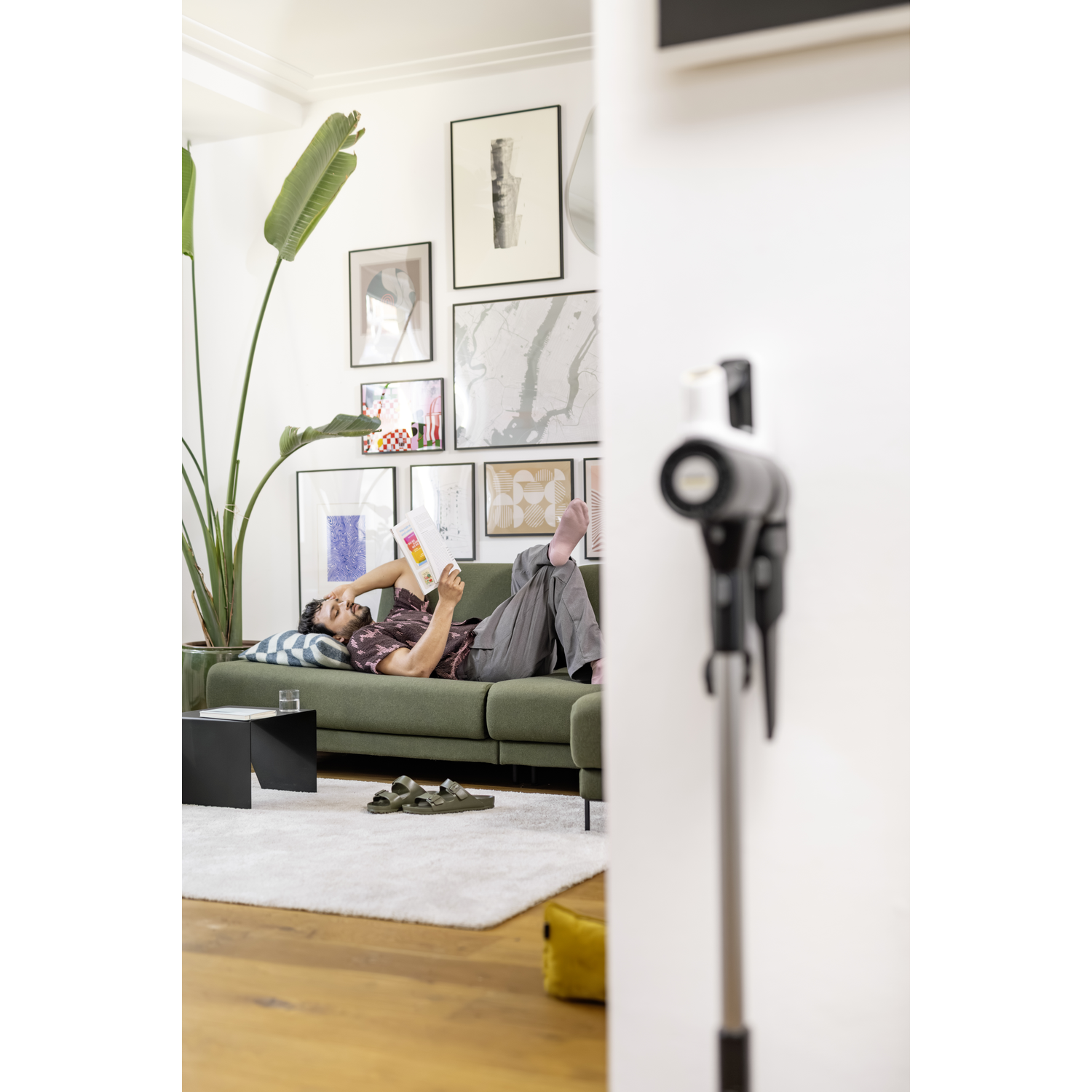 Man on a green sofa reading a magazine. A plant, art, and a vacuum cleaner are also present.