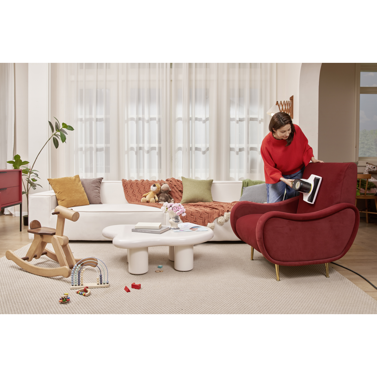 A woman in a red sweater vacuums a red armchair in a living room. A white sofa with pillows is in the background.