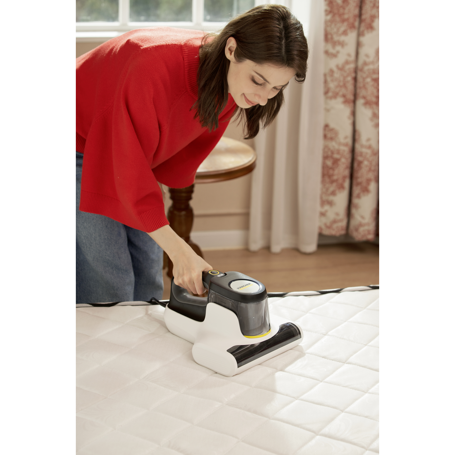 Woman in red sweater vacuums a white quilted mattress with a handheld vacuum cleaner. Background includes curtains and a round table.