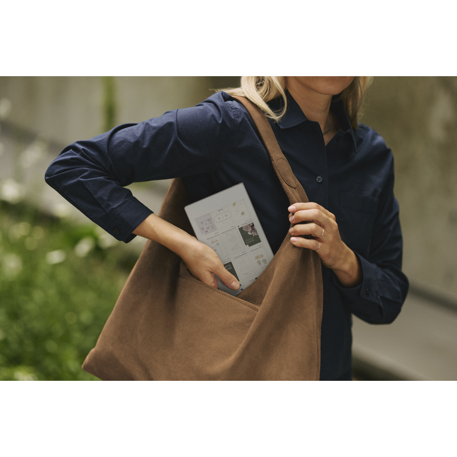 Vrouw in donkerblauw overhemd die een tablet uit een bruine tas haalt. De tas is over de schouder, met een vage groene achtergrond.
