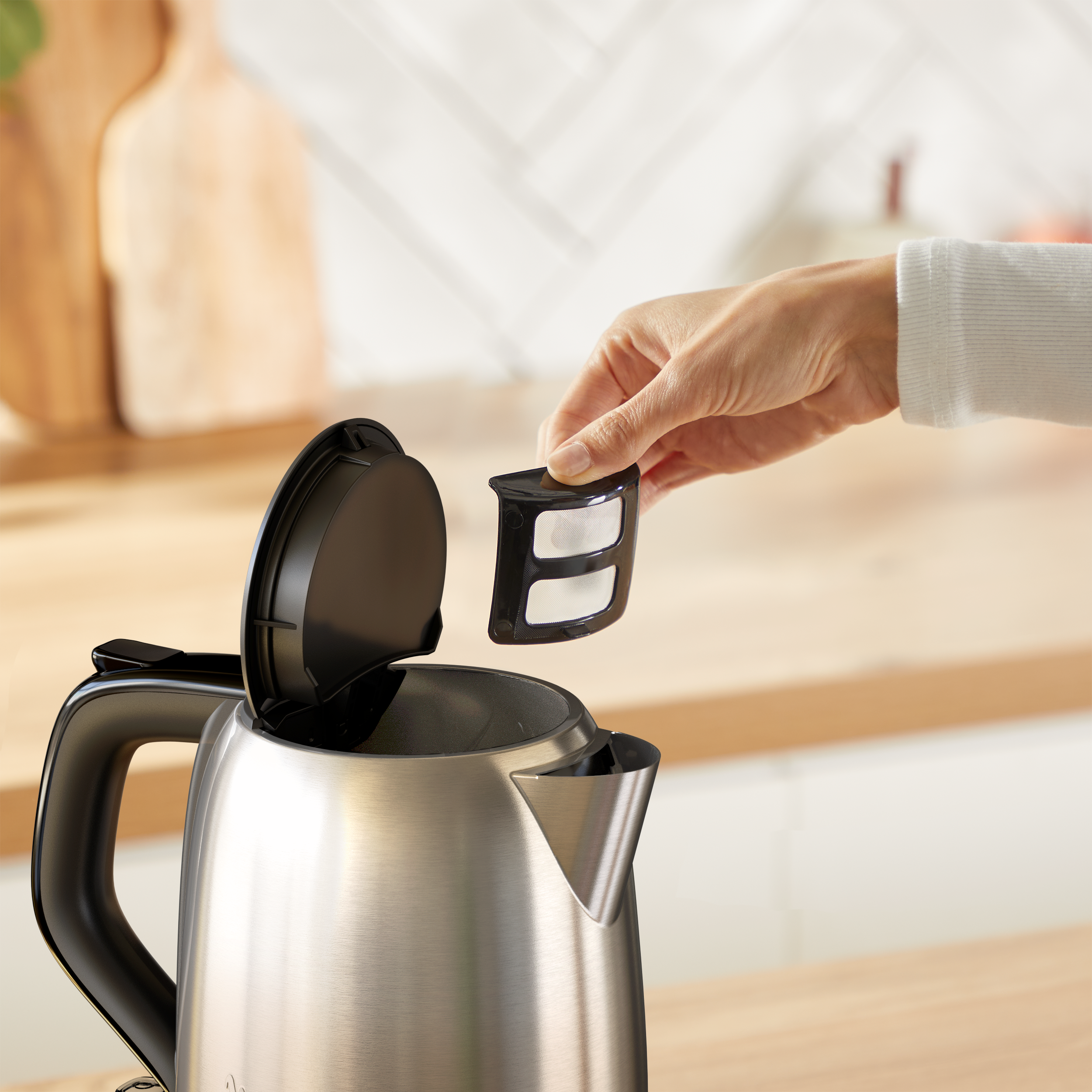 Eine Hand hält einen Teefilter über einem Wasserkocher. Der Wasserkocher ist silberfarben, mit schwarzem Griff und Deckel. Der Hintergrund ist eine Küche.