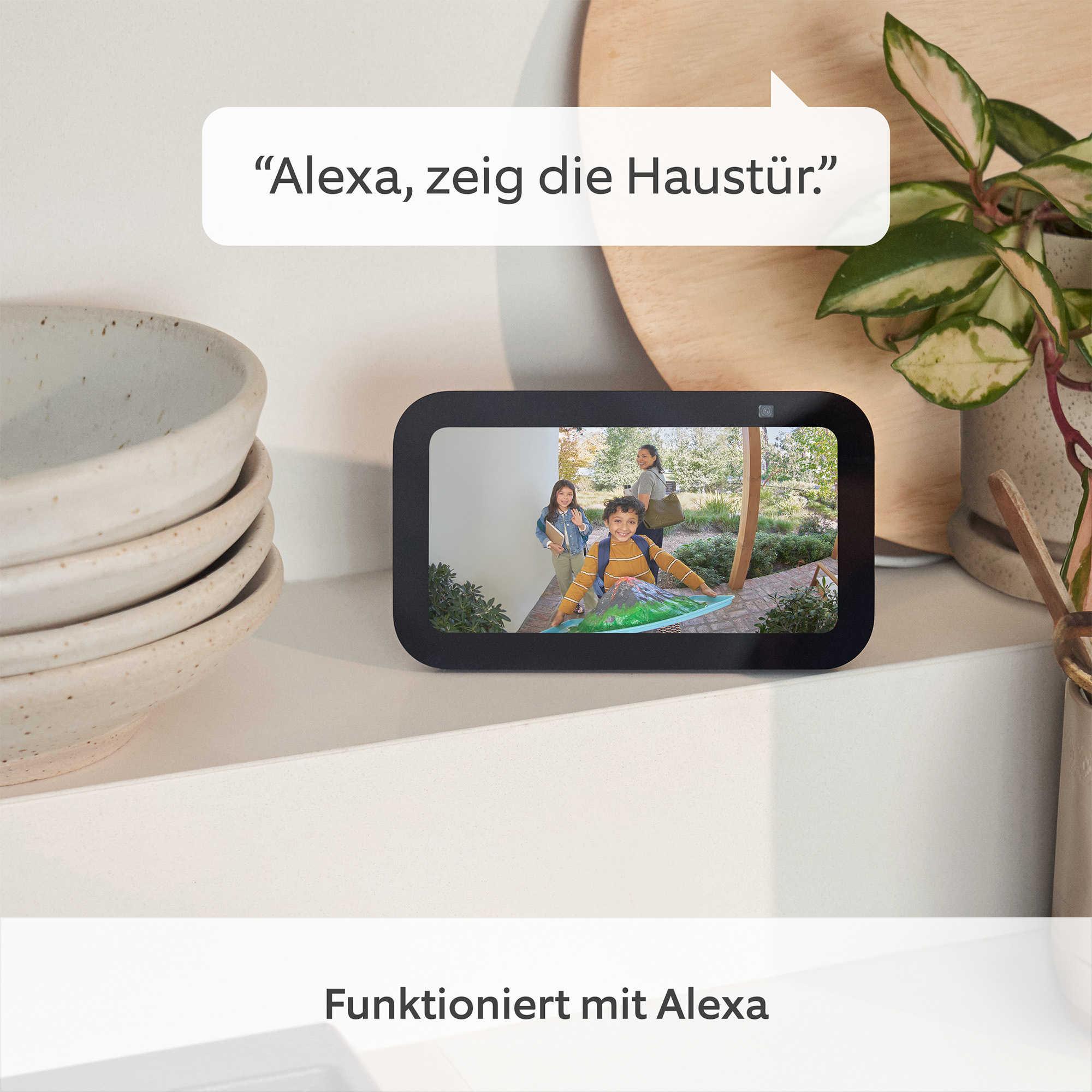 Ein Smart-Display zeigt eine Familie an der Tür. Eine Sprechblase sagt: 'Alexa, zeig die Haustür'.