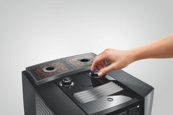 Eine Hand dreht einen Knopf an einer schwarzen Kaffeemaschine. Kaffeebohnen sind in einem Behälter oben sichtbar.