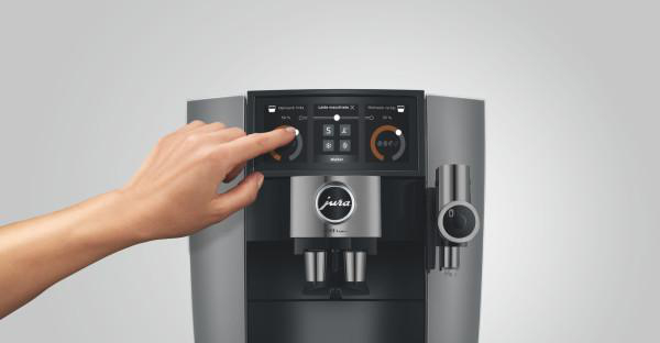 Eine Hand drückt eine Taste an einer Kaffeemaschine. Die Maschine ist grau und schwarz mit einem Touchscreen-Display.