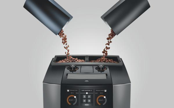 Zwei schwarze Trichter gießen Kaffeebohnen in eine Kaffeemaschine. Die Maschine hat ein Display und Drehregler und steht vor einem grauen Hintergrund.