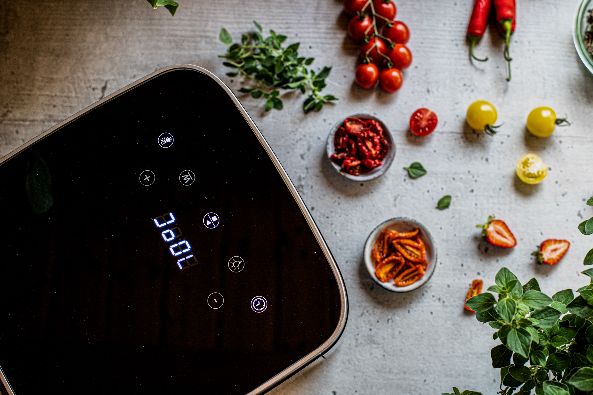 Eine Heißluftfritteuse mit Digitalanzeige, umgeben von Zutaten wie Tomaten, Chilischoten, Kräutern und sonnengetrockneten Tomaten.