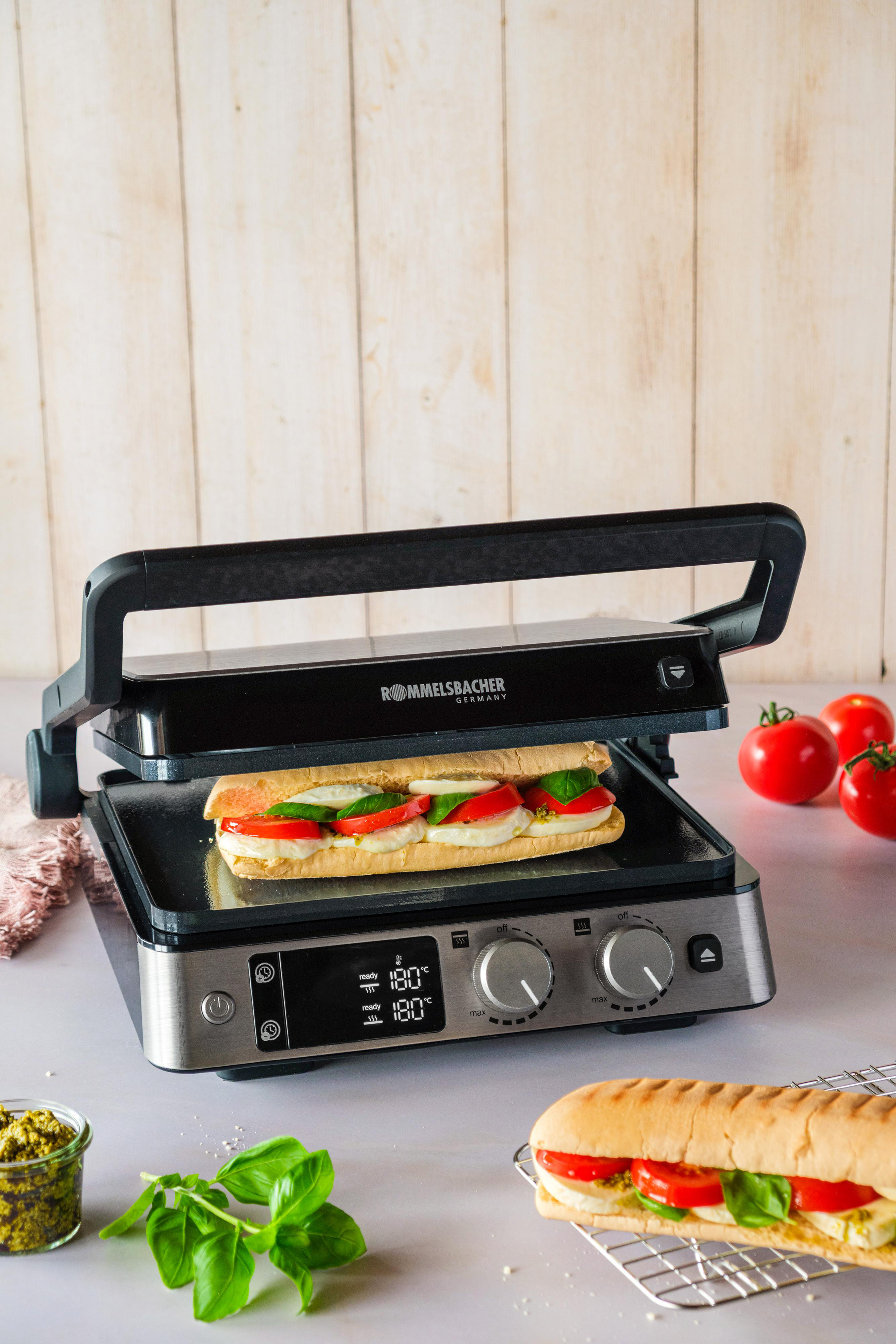 Ein Panini-Grill mit einem Sandwich. Tomaten, Basilikum und Pesto sind daneben. Holziger Hintergrund und ein Sandwich auf einem Rost.