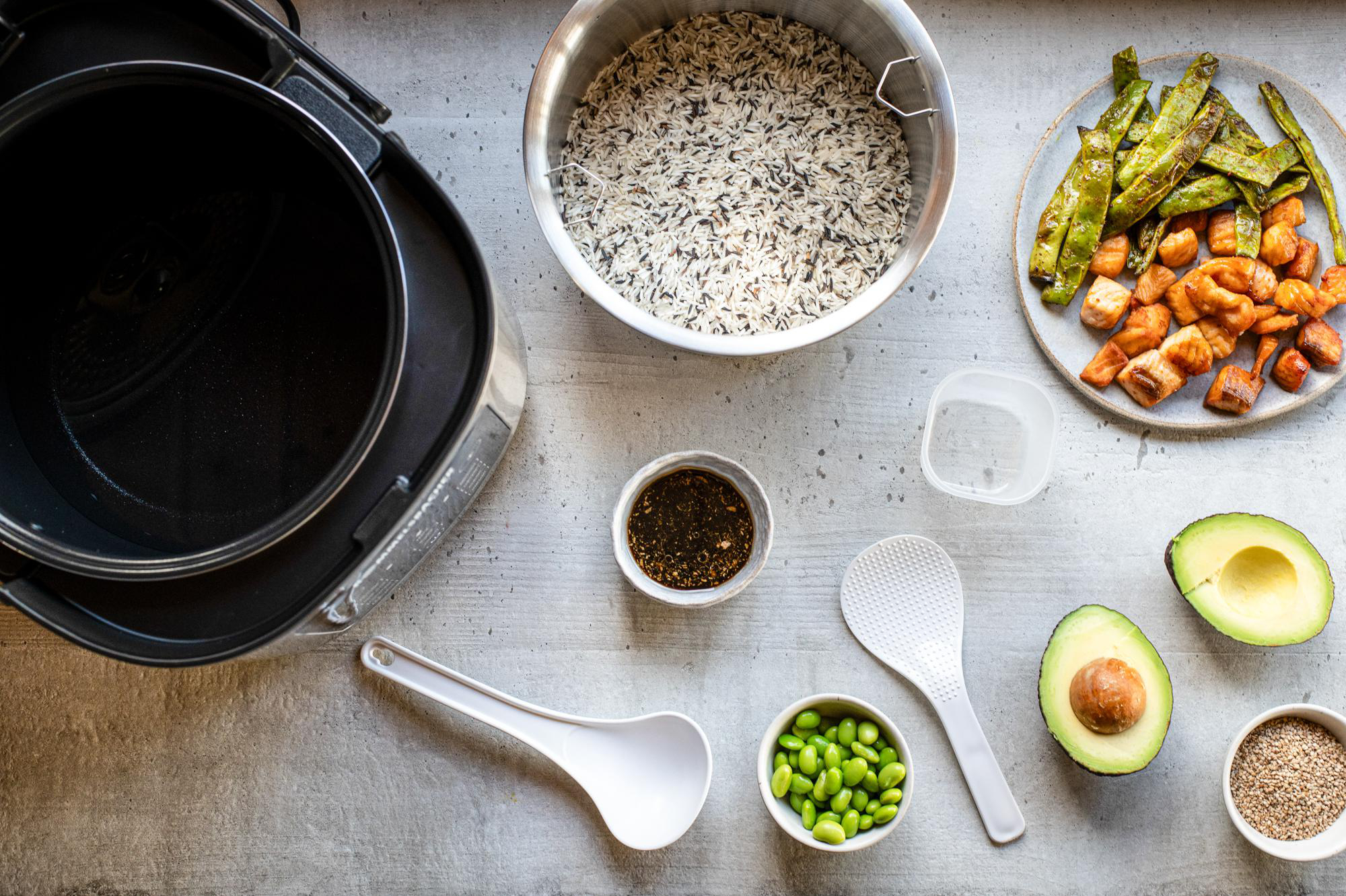 Draufsicht auf verschiedene Lebensmittel: Reis, Avocado, Lachs, grüne Bohnen, Edamame und Saucen, neben einem Reiskocher und Utensilien.
