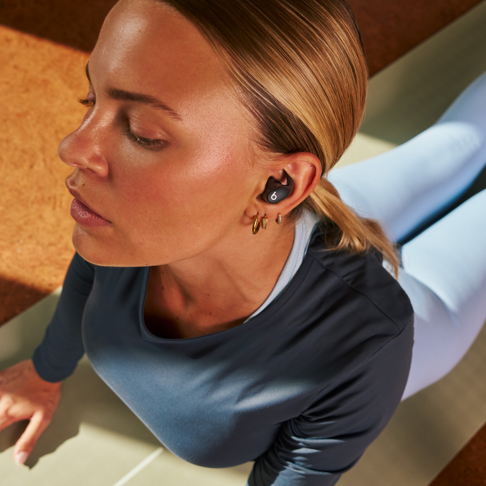 Frau in Yoga-Pose mit Beats-Kopfhörern. Trägt blaue Leggings und ein Top. Hintergrund ist eine Korkoberfläche und eine grüne Matte.
