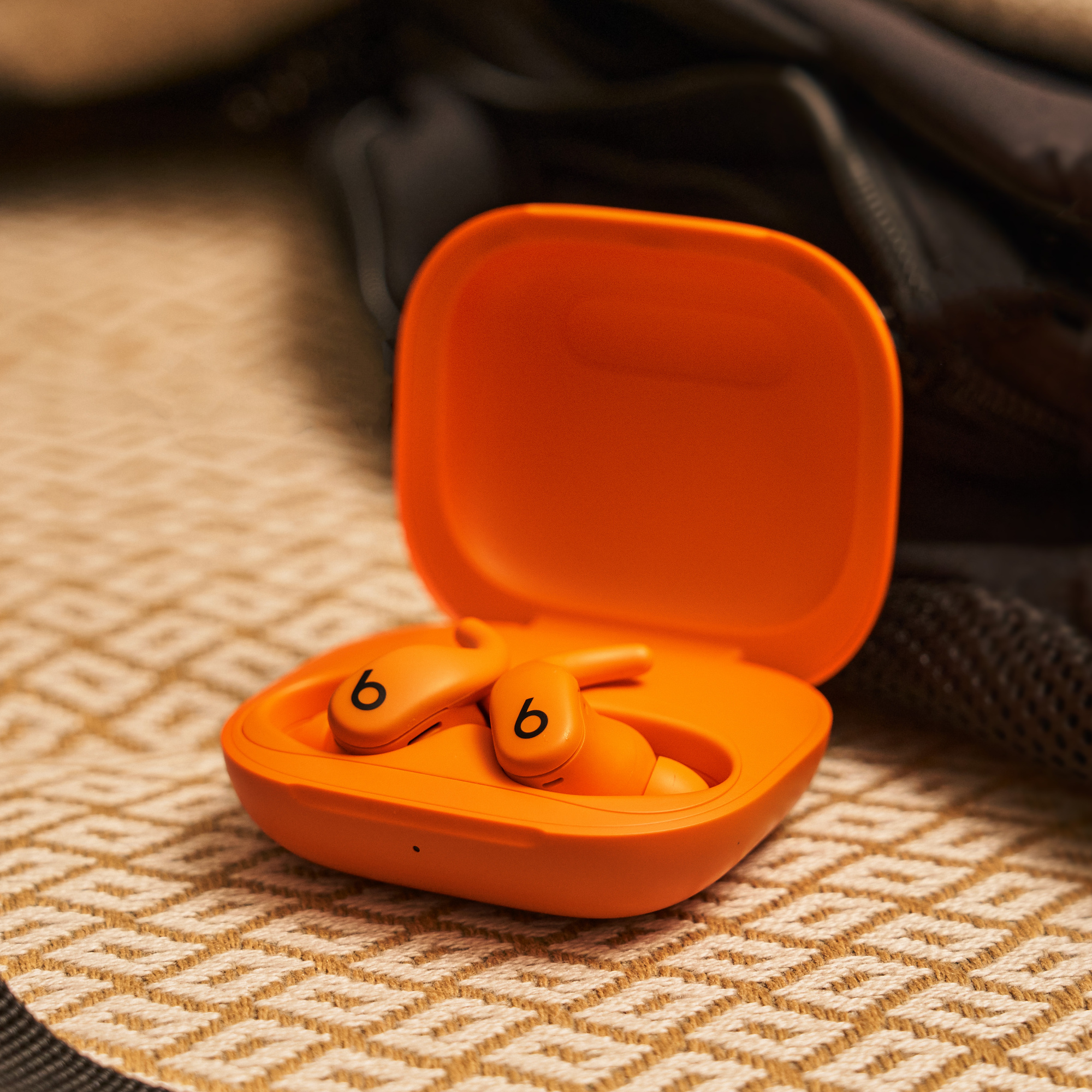 Helle orangefarbene Beats-Ohrhörer in einem geöffneten Ladekoffer auf einer gemusterten Oberfläche, ein dunkler Rucksack im Hintergrund.