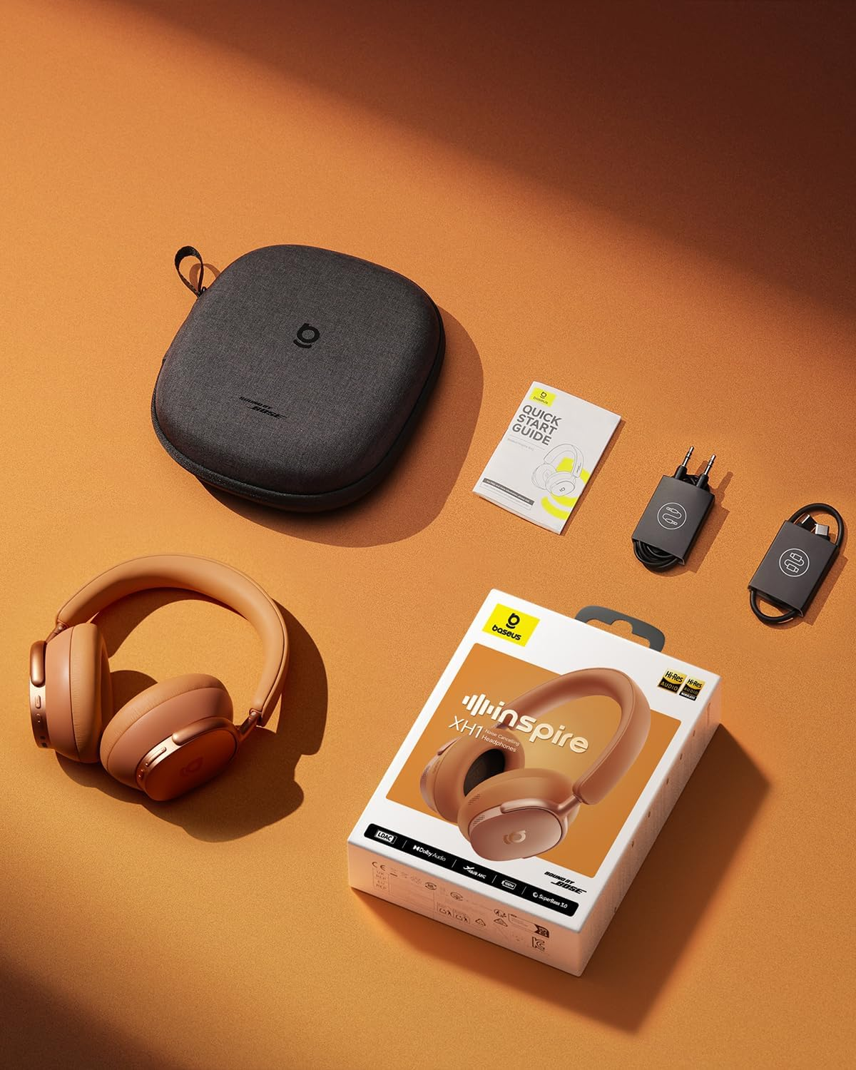 Eine Draufsicht zeigt Kopfhörer, Etui, Schachtel und Zubehör auf einer orangefarbenen Oberfläche. Die Kopfhörer sind braun, die Box ist weiß.