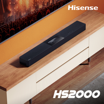 Una barra de sonido negra encima de un mueble de madera. El fondo muestra una pantalla de TV y una multitud en un concierto.