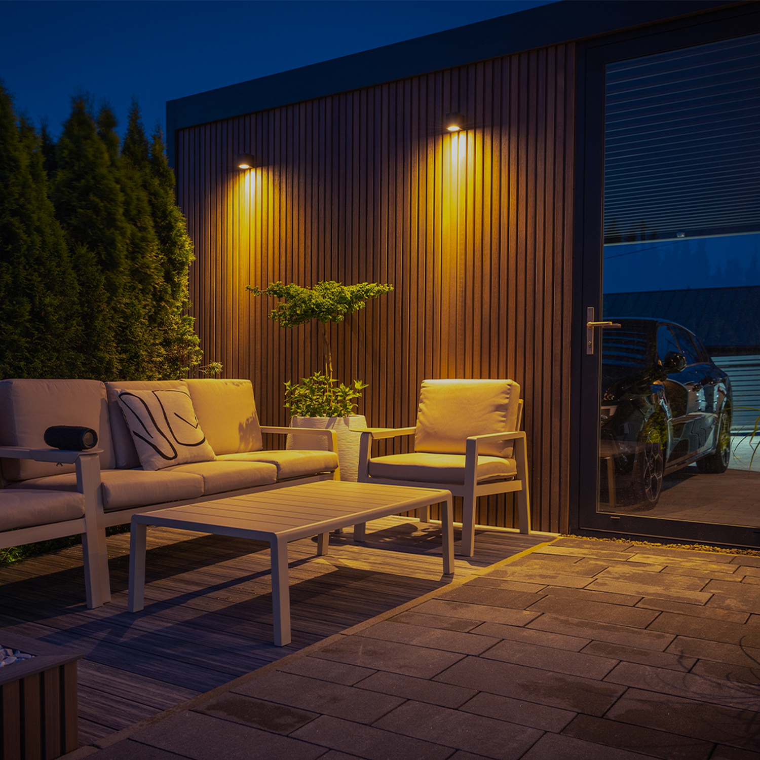 Zona de estar al aire libre por la noche. Sofá, mesa y silla beige. Pared de madera con luces. Coche visible por puerta de cristal.