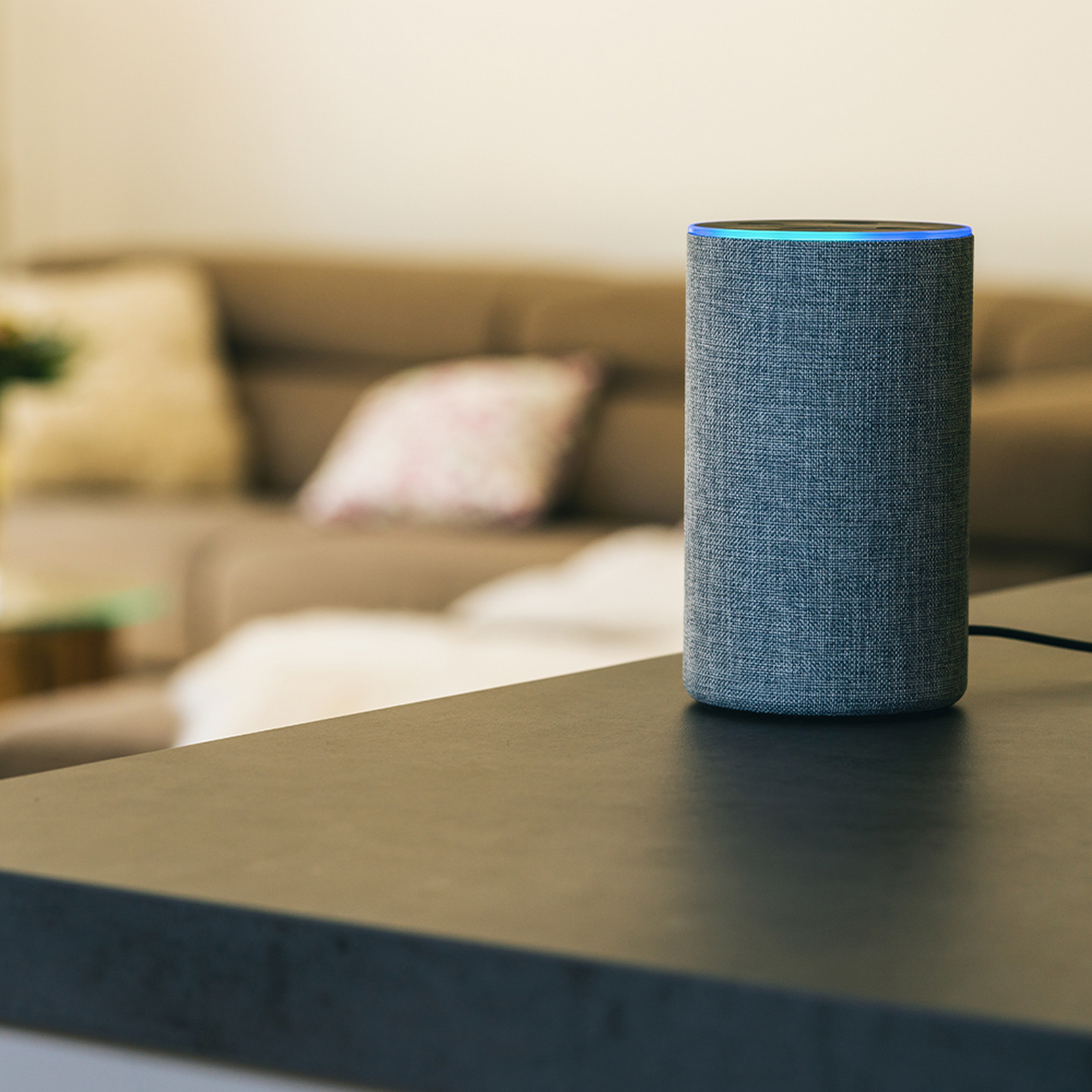 Un altavoz inteligente cilíndrico con una parte superior azul brillante está sobre una superficie negra. Un sofá y almohadas están borrosos en el fondo.