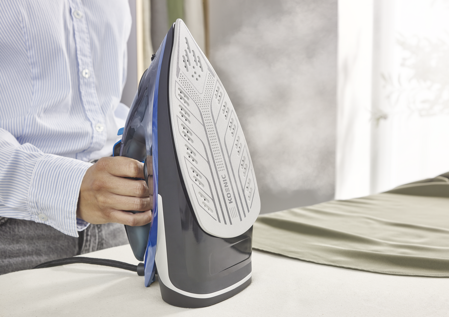 Una persona planchando con una plancha de vapor azul y negra. El vapor sale de la plancha. Tela verde en la tabla de planchar.