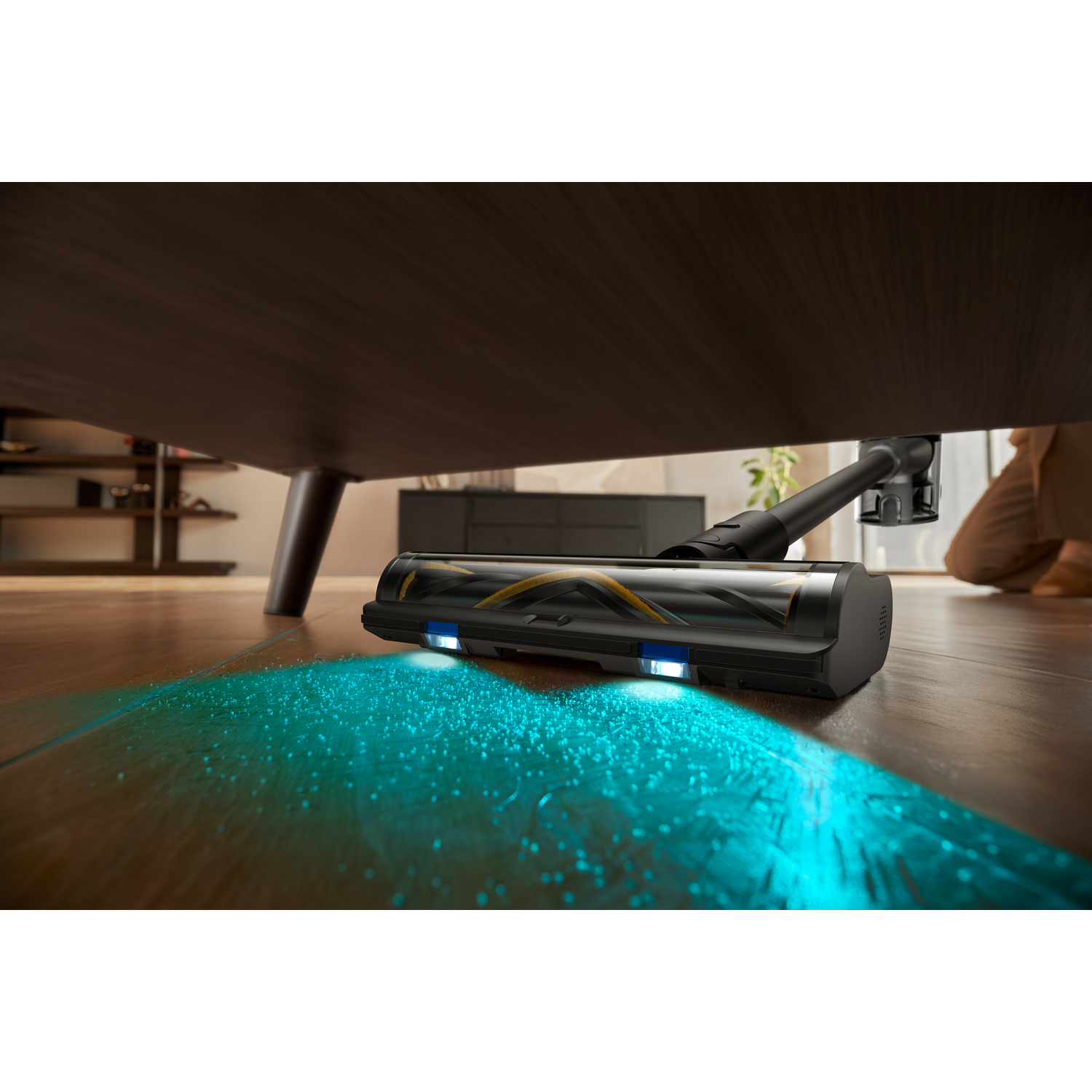 Een stofzuiger maakt schoon onder een donkere houten tafel. Een persoon is op de achtergrond. De stofzuiger geeft blauw licht af.