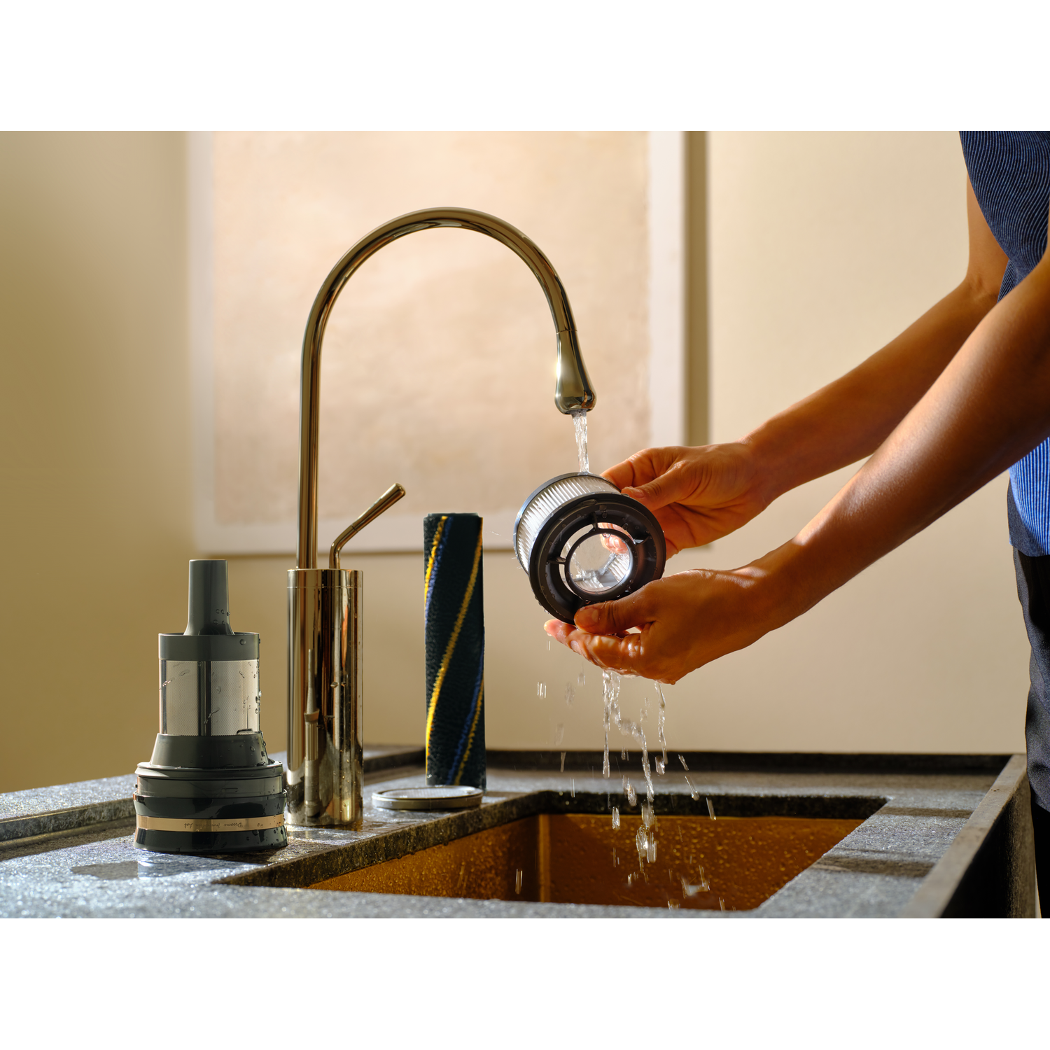 Een persoon wast een stofzuigerfilter onder een gouden kraan. Andere onderdelen van de reiniger liggen in de wasbak. Water stroomt.