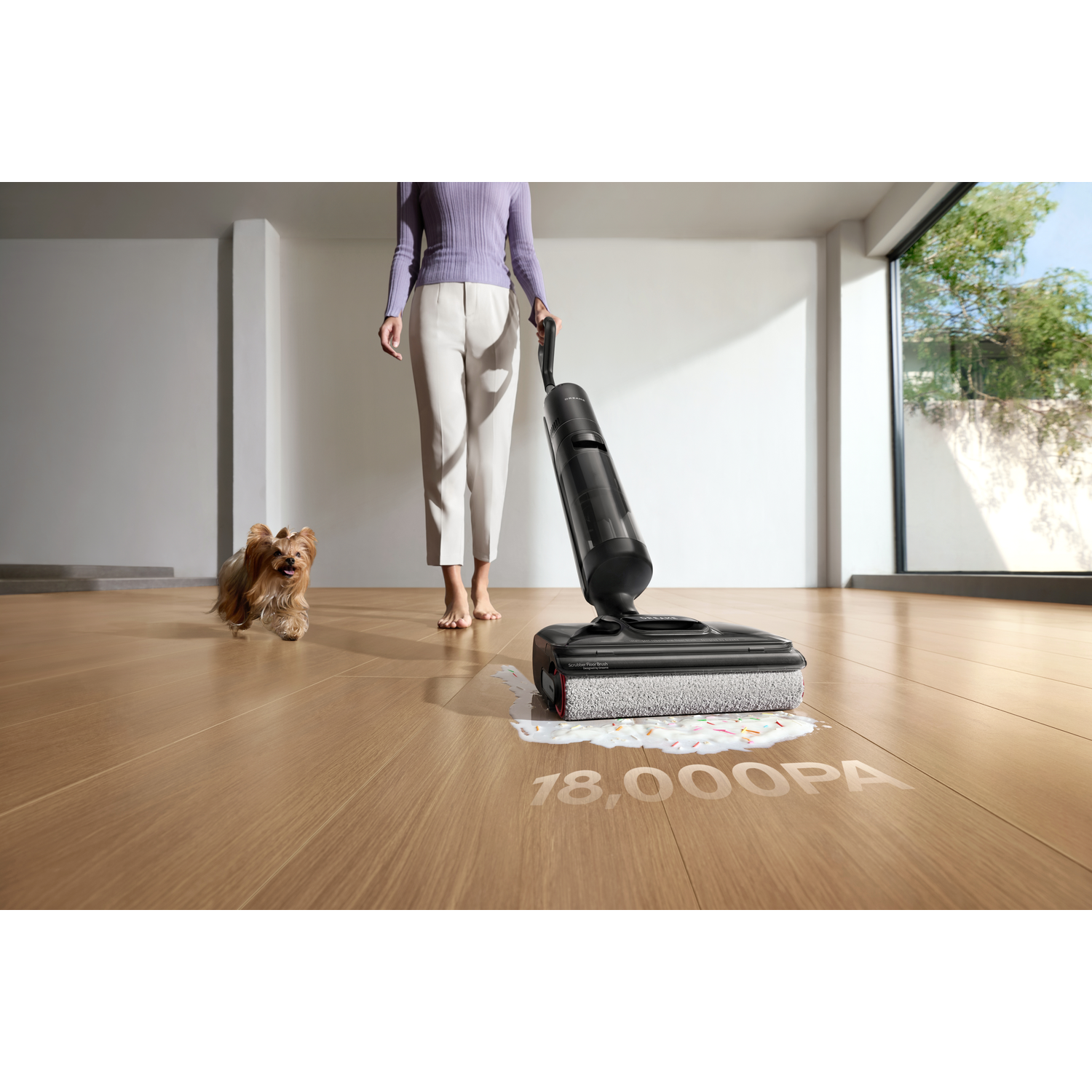 Une femme utilise un aspirateur sur un sol en bois. Un petit chien court au premier plan. Le texte sur le sol dit '18,000PA'.