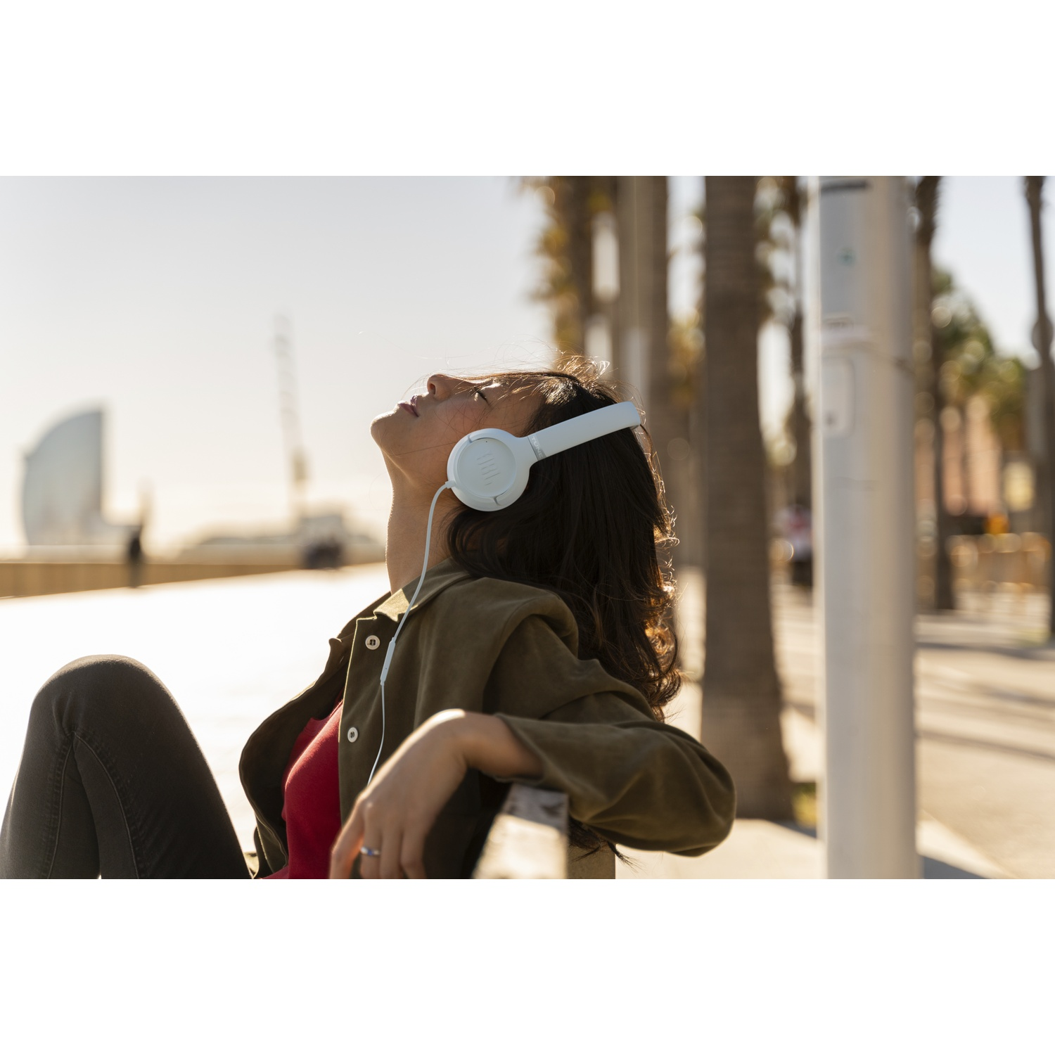 A woman wearing white headphones sits on a bench, looking up at the sun. Palm trees and a building are in the background.