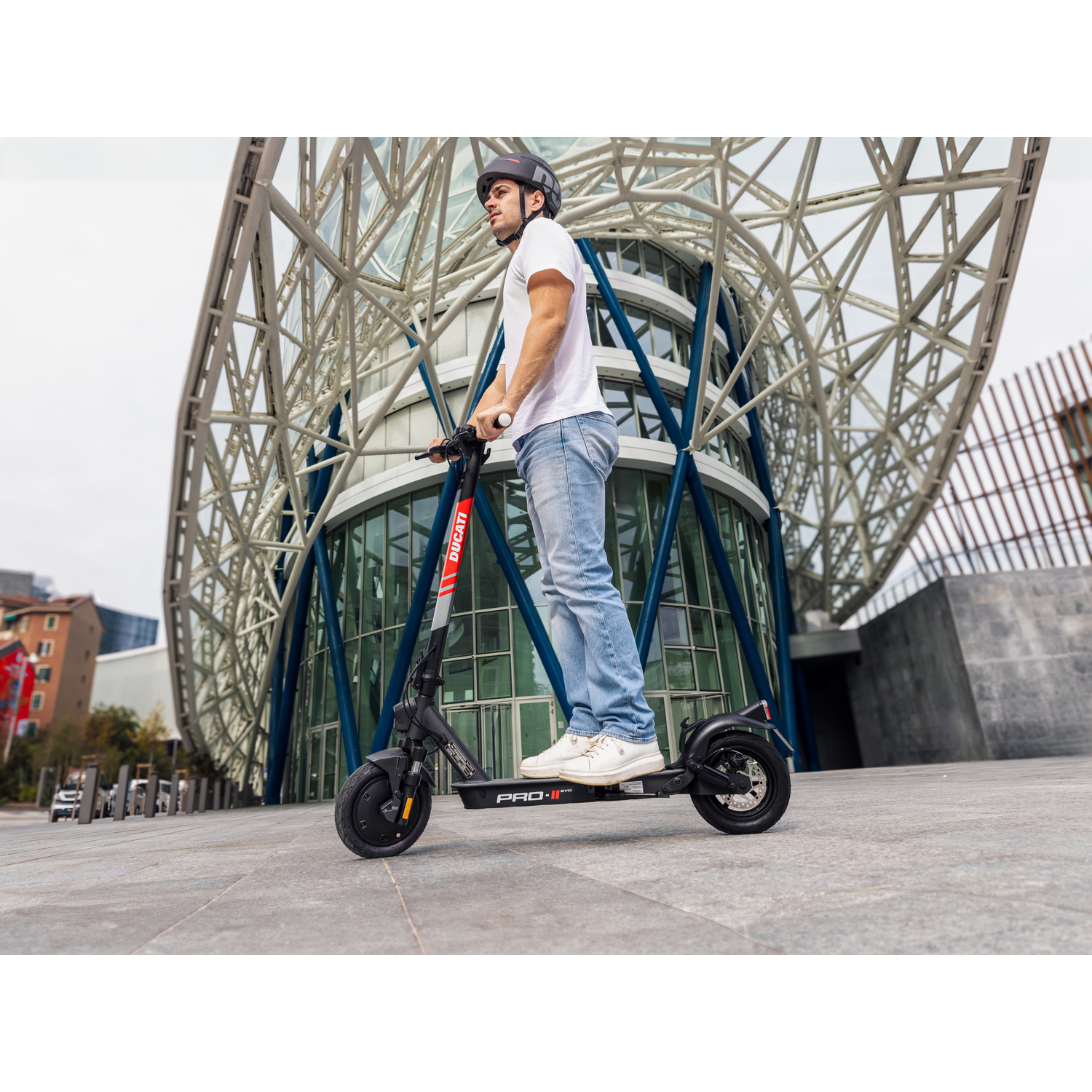 Een man met een helm rijdt op een zwarte Ducati scooter op een geplaveid gebied voor een modern gebouw.
