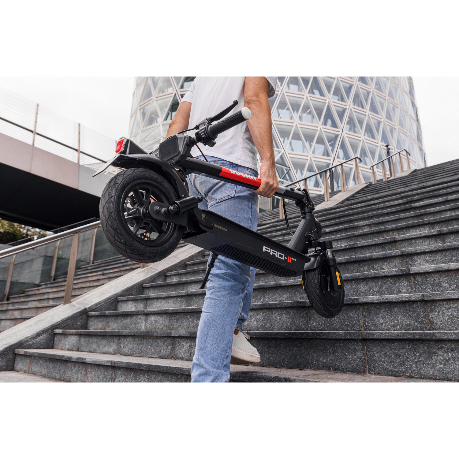 Een persoon draagt een zwarte elektrische scooter de trap op. De achtergrond toont een modern gebouw en een bewolkte lucht.
