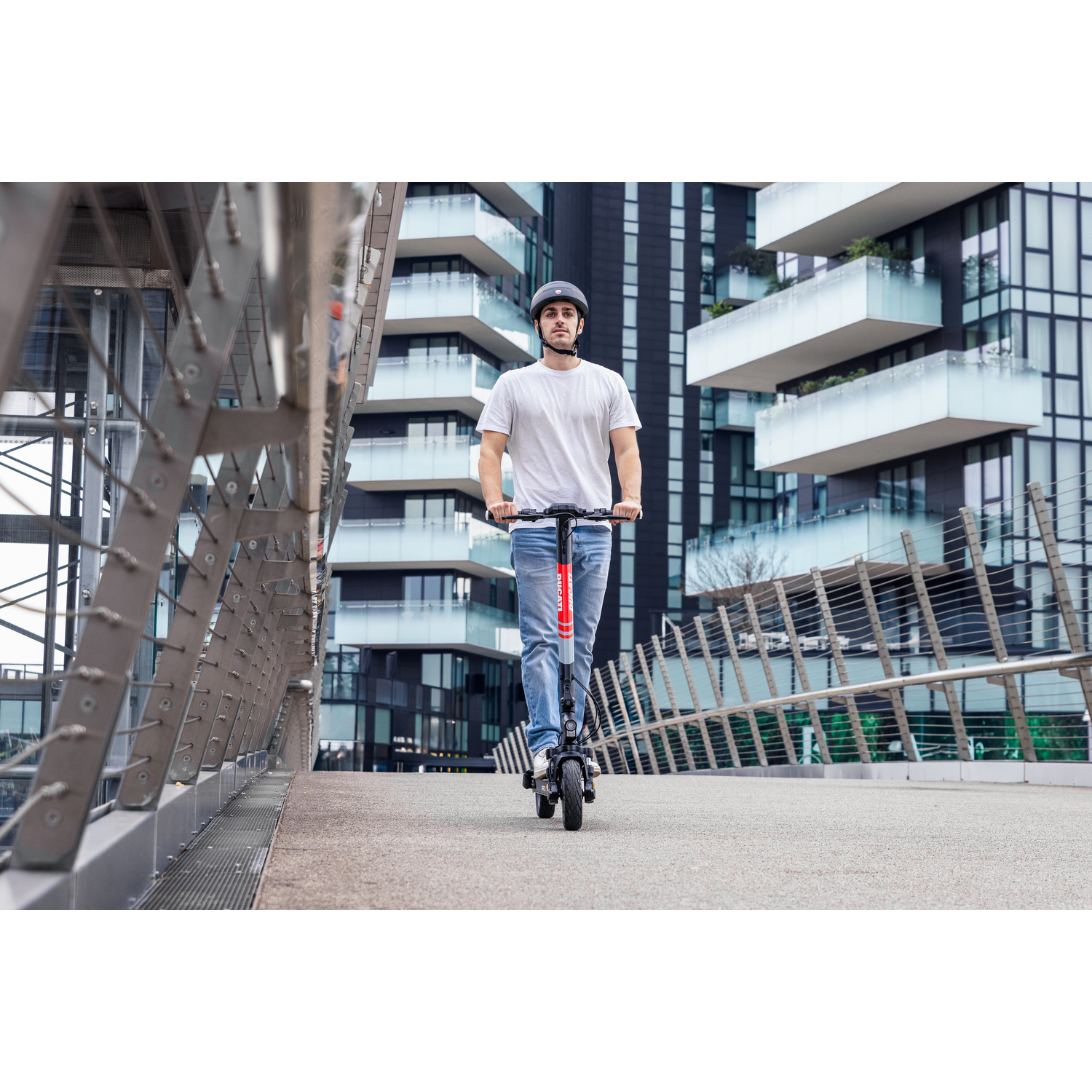 Een man rijdt op een elektrische scooter op een brug. Hij draagt een helm, een wit shirt en een spijkerbroek. Gebouwen op de achtergrond.