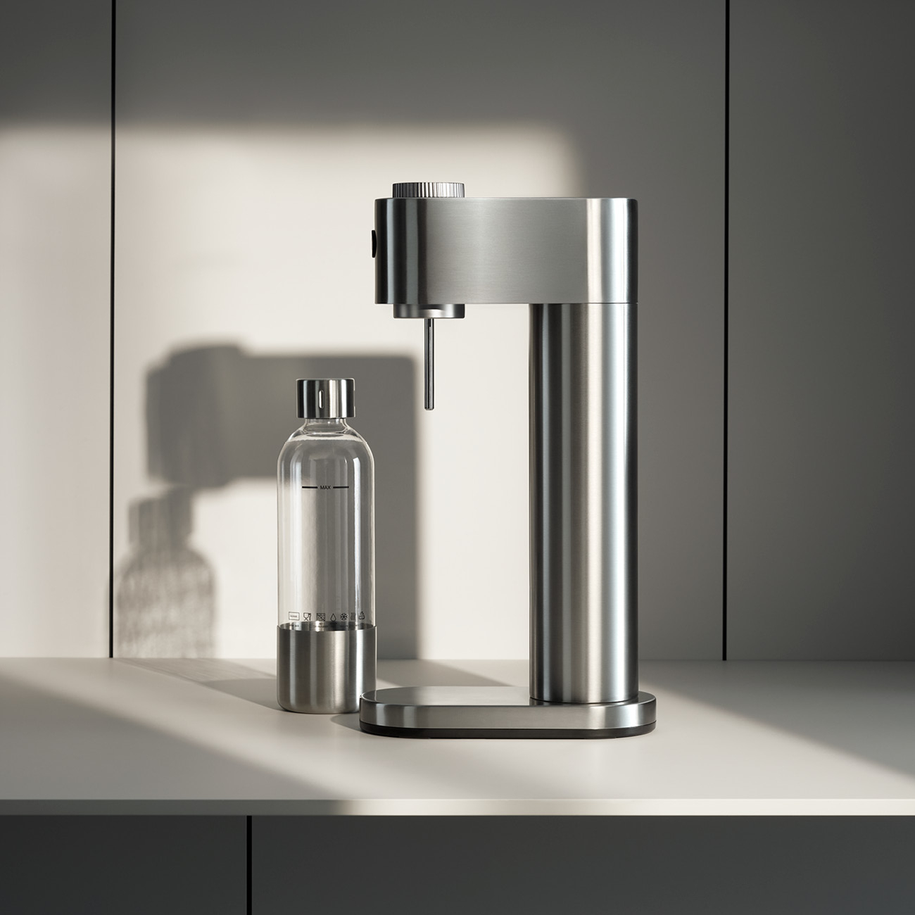 Ein Soda-Maker und eine Glasflasche aus Edelstahl stehen auf einer weißen Oberfläche. Im Hintergrund eine Wand mit Schatten.