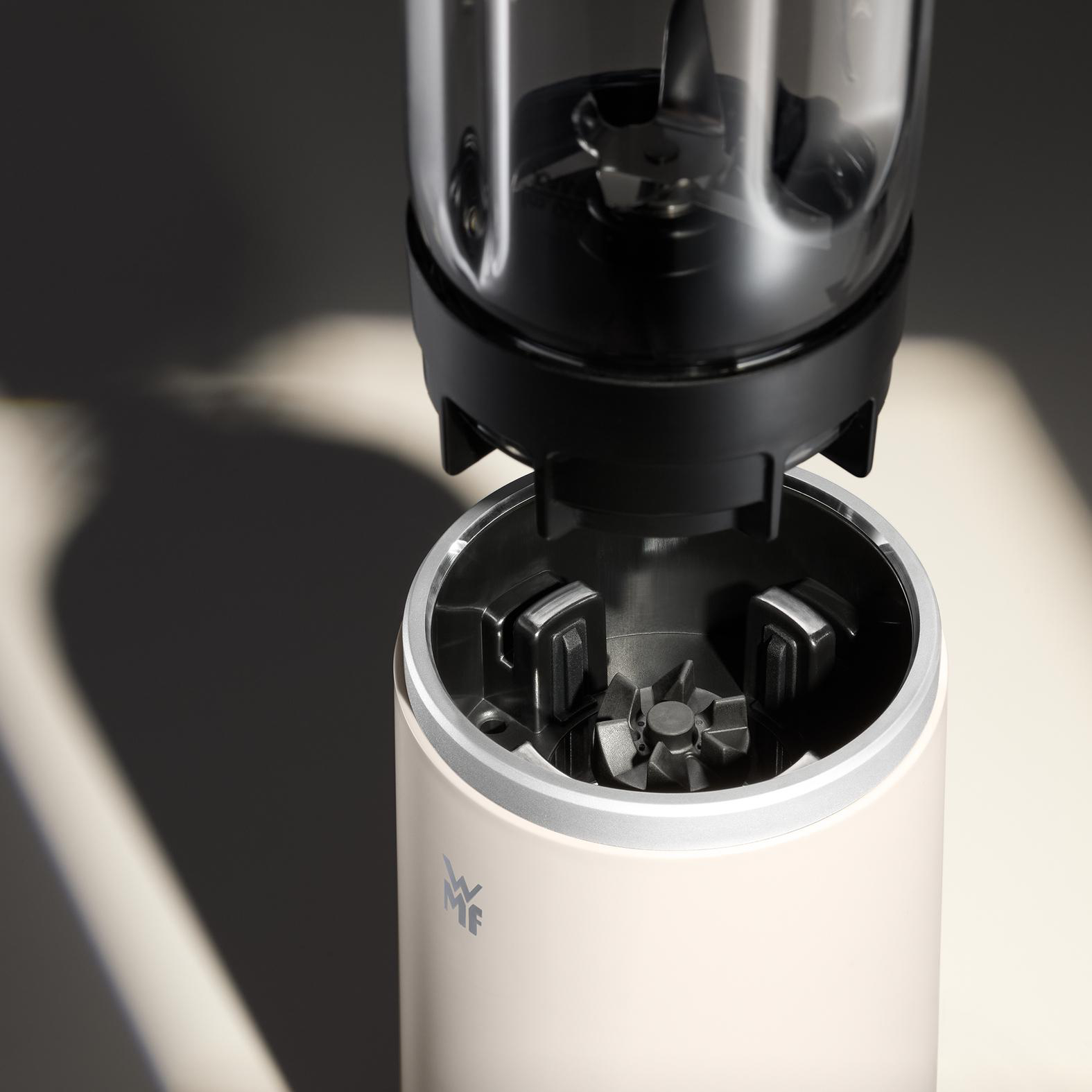 Ein Mixer, zerlegt. Der obere Teil mit Klingen befindet sich über der Basis. Die Basis ist hell und hat einen silbernen Rand.