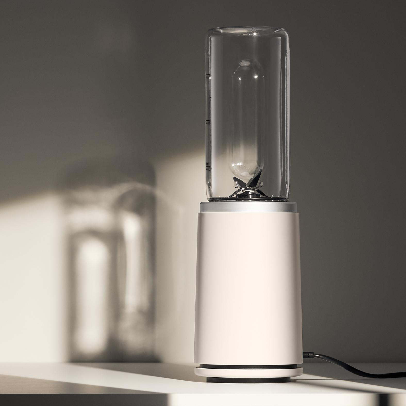 Ein Mixer mit weißem Sockel und klarem Glaskopf. Ein Schatten des Mixers ist an der Wand dahinter.