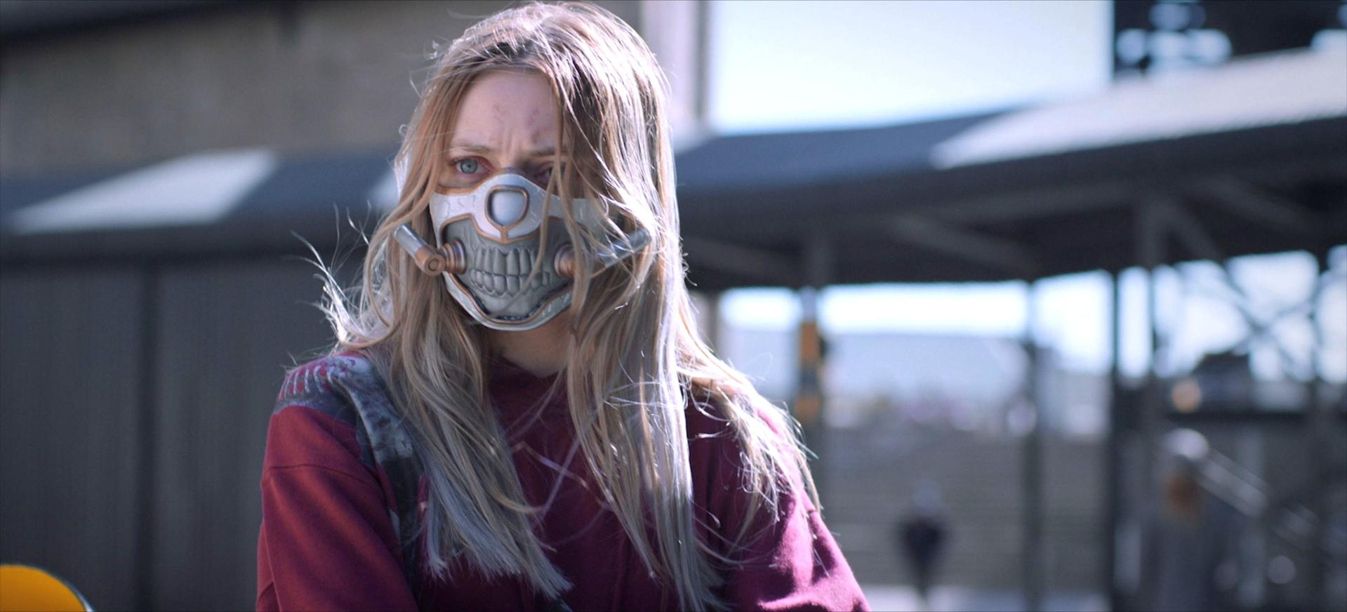 Eine Frau mit langen blonden Haaren trägt eine Maske, die Zähne darstellt. Sie steht vor einem Gebäude und trägt ein rotes Hemd.