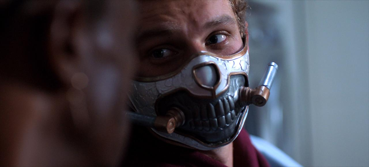 Ein Mann trägt eine Maske mit schädelartigem Design. Details umfassen Schläuche, Metall und eine Nahaufnahme des Gesichts.