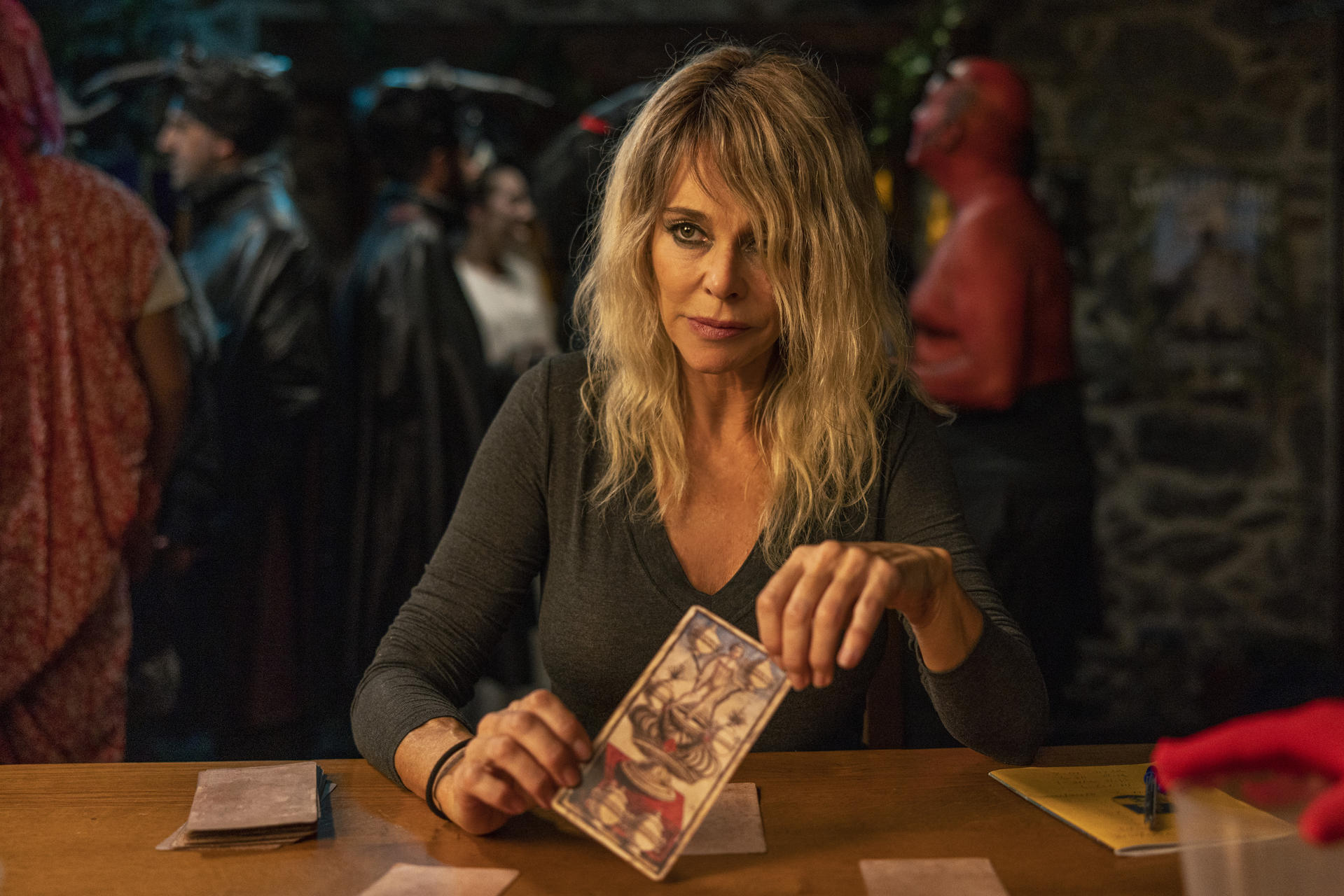 Eine Frau mit blondem Haar hält eine Tarotkarte. Sie sitzt an einem Tisch mit anderen Karten. Im Hintergrund sind weitere Personen zu sehen.