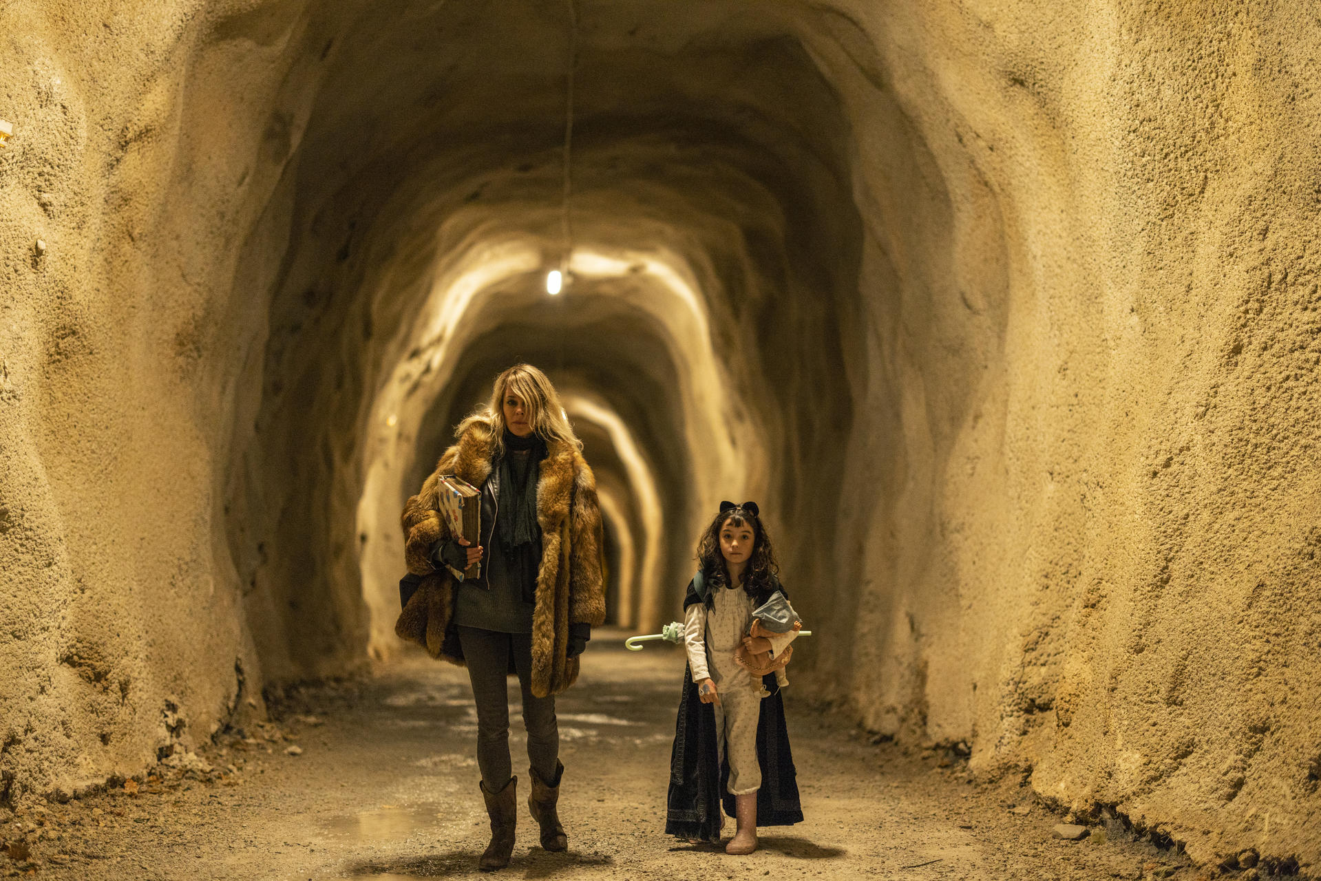 Eine Frau und ein Mädchen stehen in einem Tunnel. Die Frau trägt einen Pelzmantel und Stiefel, das Mädchen hat einen Umhang und Ohren.