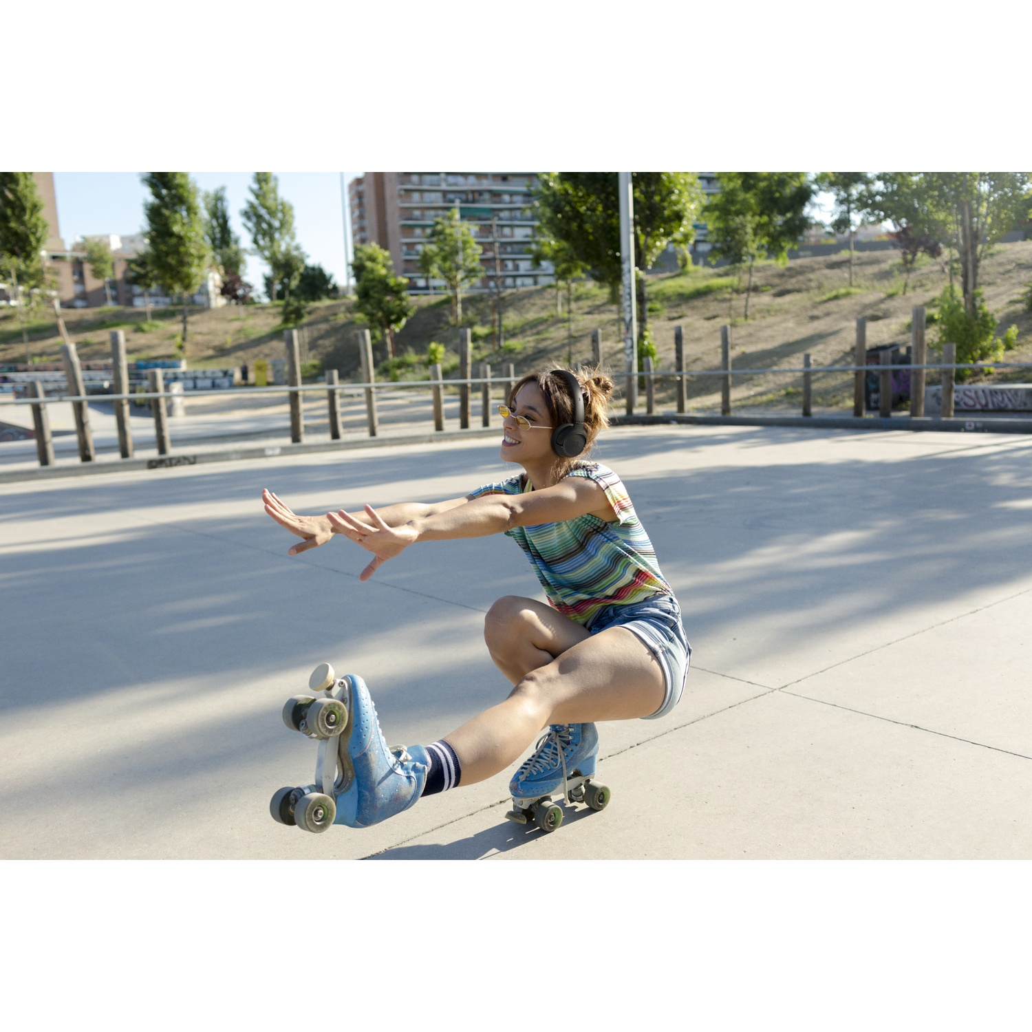 A woman on roller skates balances. She wears headphones, sunglasses, and shorts. The background shows a park with trees and buildings.
