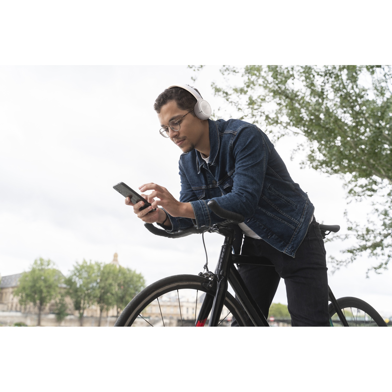 Un homme avec des lunettes et un casque utilise son téléphone en s'appuyant sur un vélo noir. Arbres et bâtiment en arrière-plan.