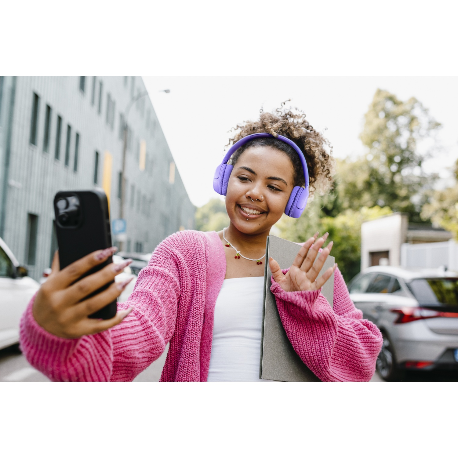 Une jeune femme sourit, tenant un téléphone pour un selfie. Elle porte un casque violet et un pull rose. Des voitures et un bâtiment en arrière-plan.