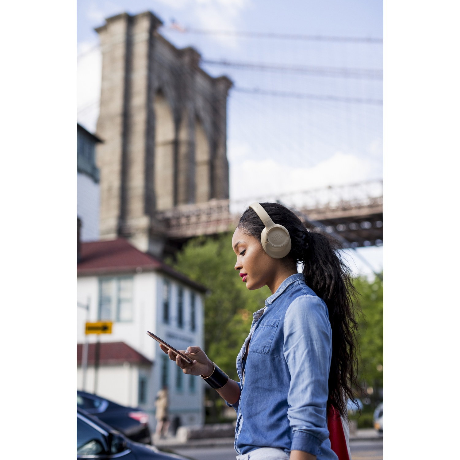 Een vrouw met een koptelefoon en een spijkerblouse kijkt naar haar telefoon. De Brooklyn Bridge staat op de achtergrond.