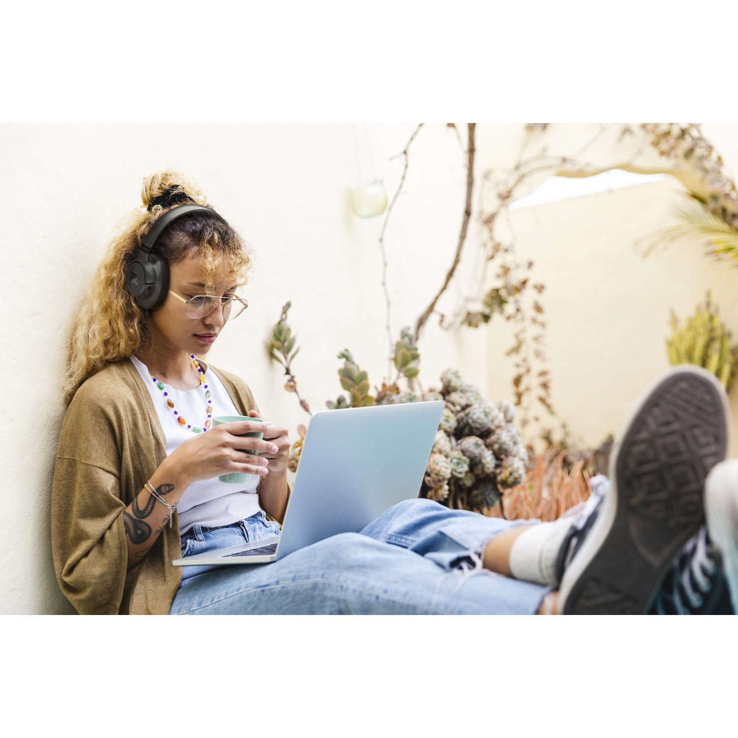 Une femme avec un casque est assise avec un ordinateur portable, tenant une tasse. Elle porte des lunettes, un cardigan et un jean, avec un tatouage. Des plantes en arrière-plan.
