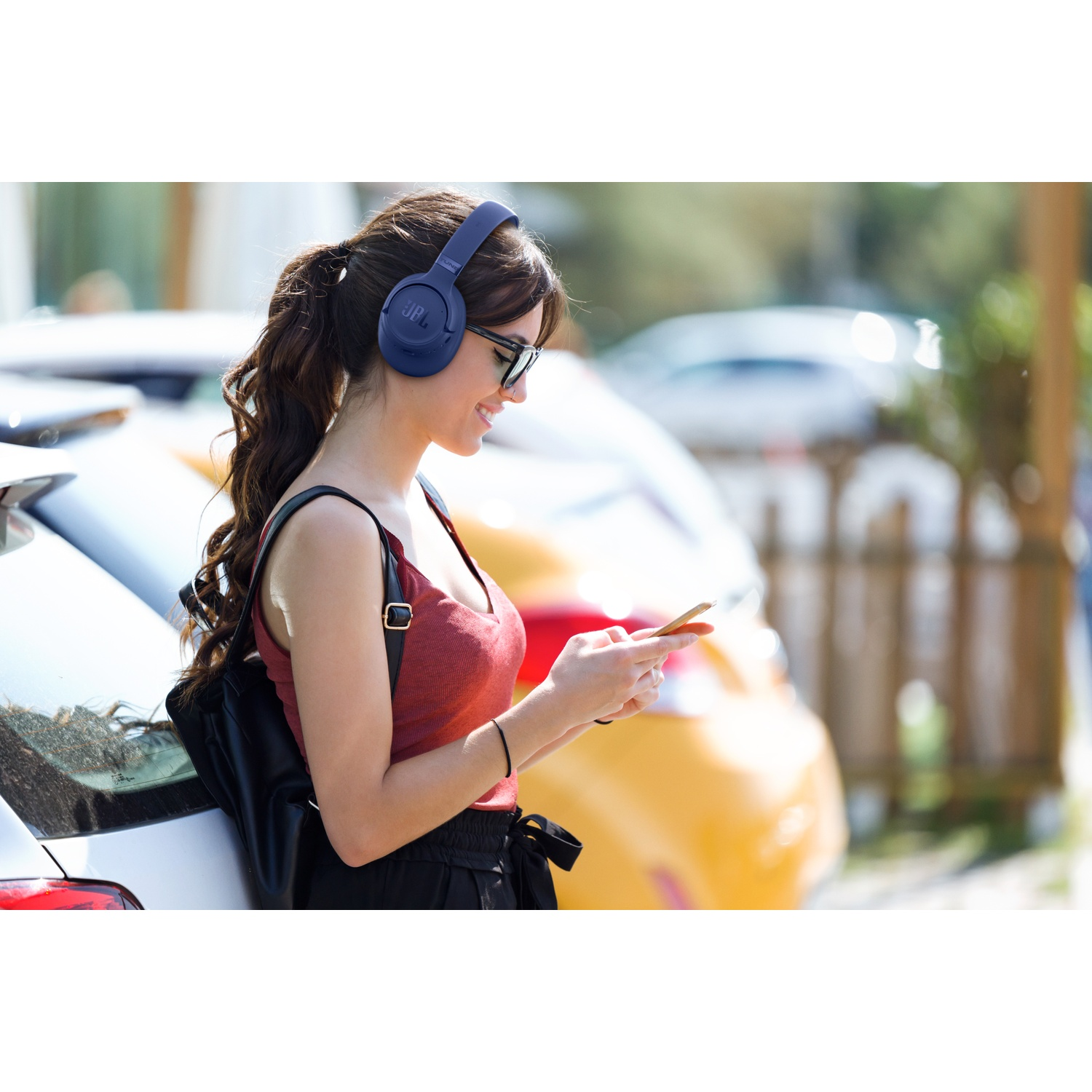 Une femme en haut rouge porte un casque, des lunettes de soleil et regarde son téléphone près de voitures garées. Elle a un sac à dos.