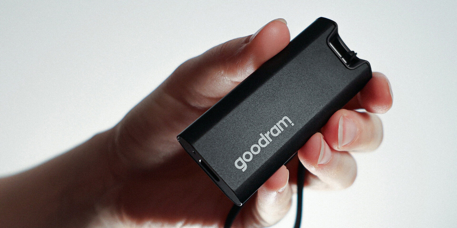 Dłoń trzyma czarny pendrive z napisem 'goodram'. Białe tło. Do pendrive'a podłączony jest kabel.