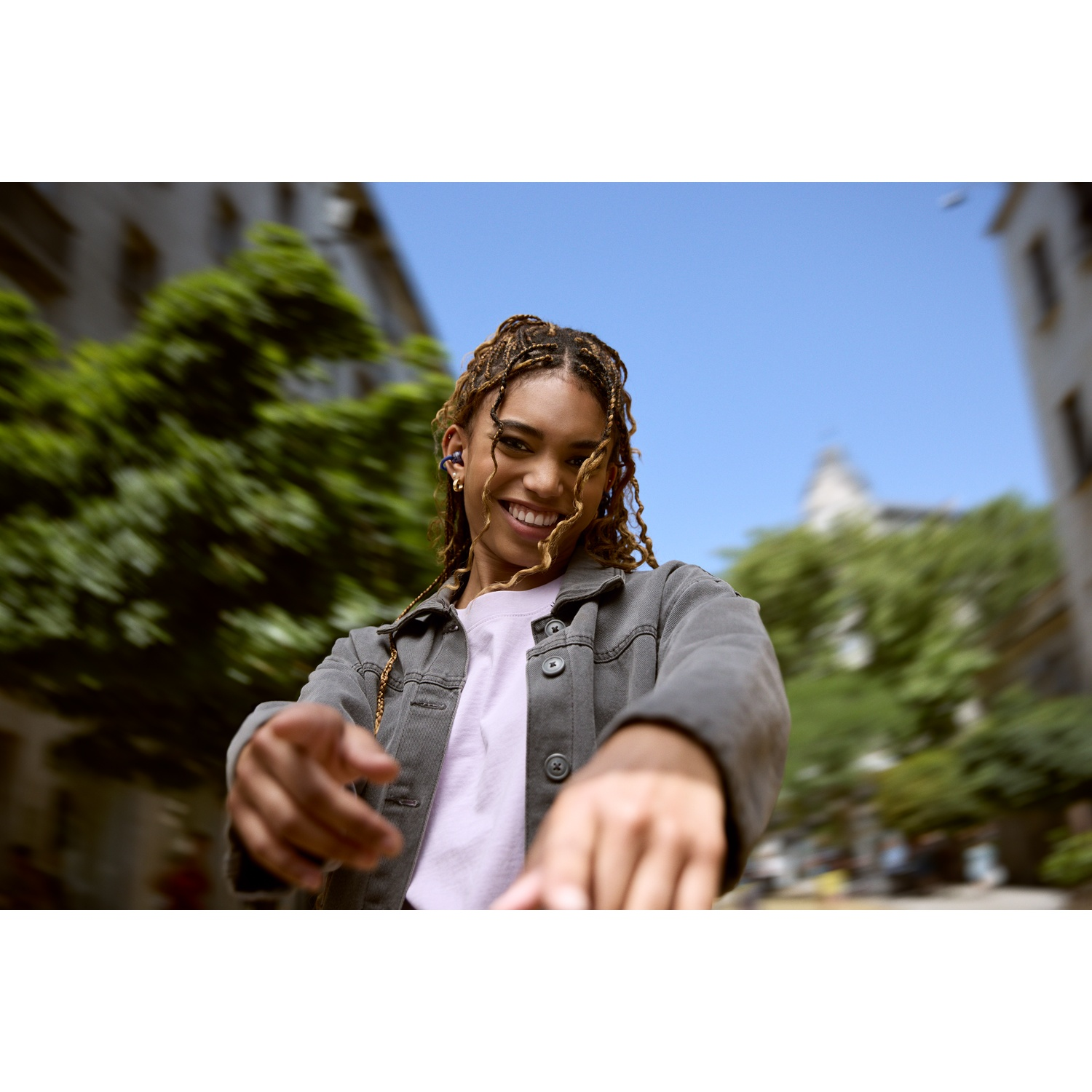 Une femme souriante aux cheveux tressés pointe vers le spectateur. Elle porte une veste grise et une chemise rose, avec un fond de ciel bleu.