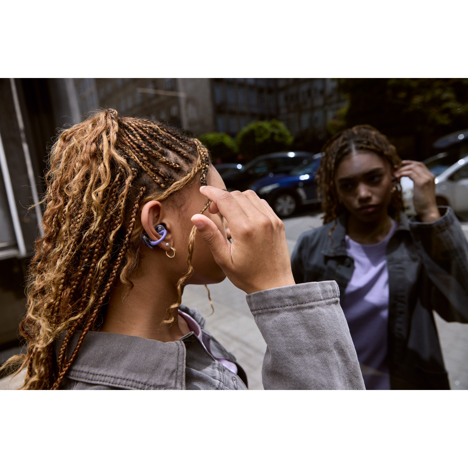 Deux femmes aux cheveux tressés à l'extérieur. L'une touche son oreille avec un casque, l'autre ajuste sa boucle d'oreille. Des voitures en arrière-plan.