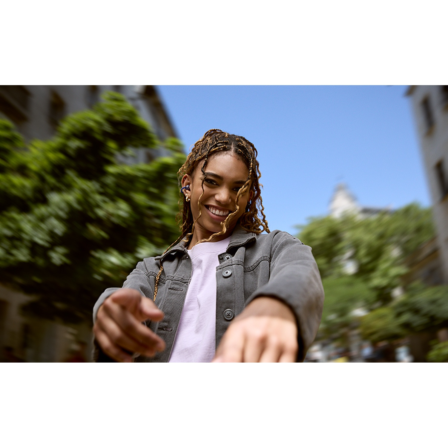 Une femme souriante aux cheveux tressés pointe vers le spectateur. Elle porte une veste grise, une chemise blanche et des boucles d'oreilles. Bâtiments et arbres en arrière-plan.