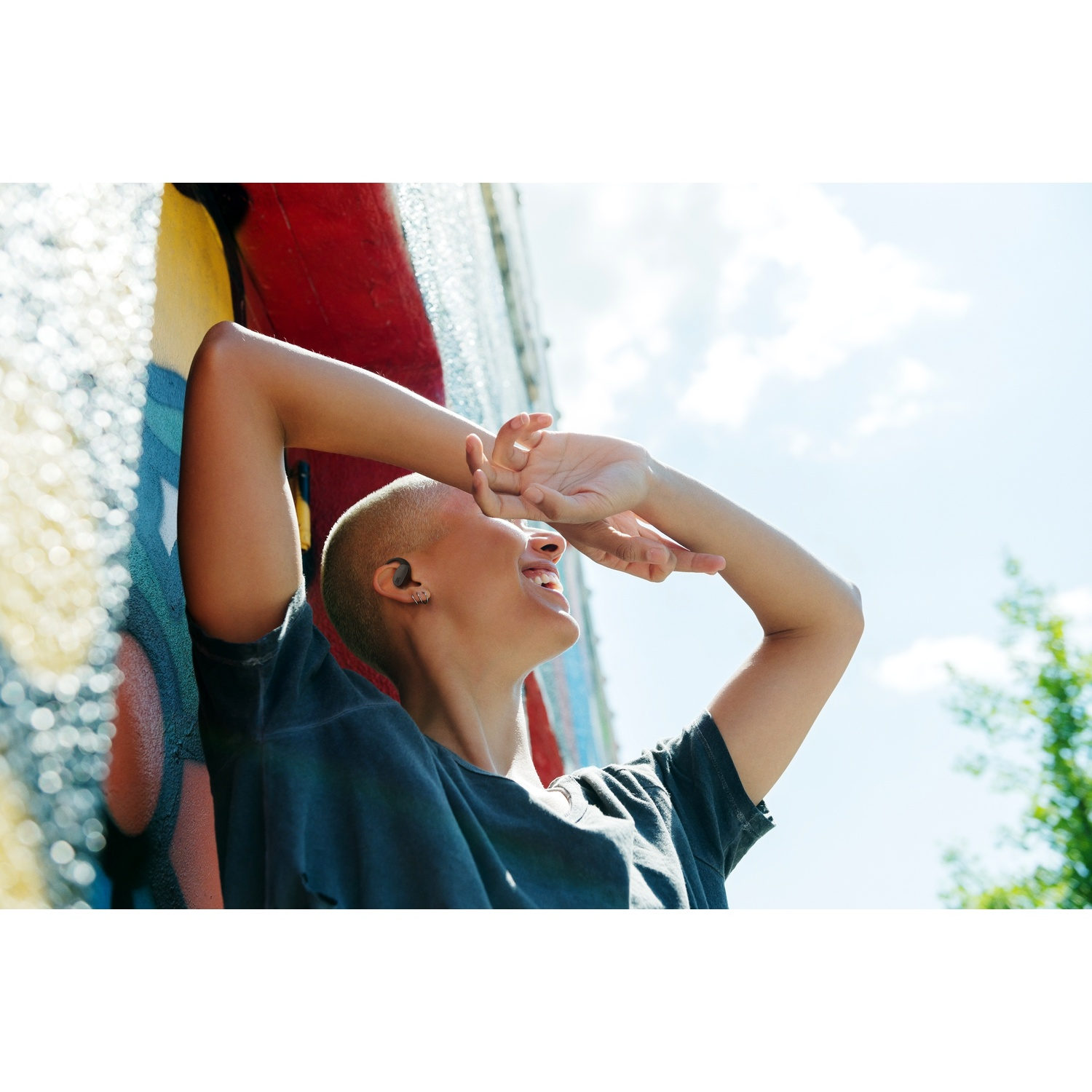 Une personne aux cheveux courts sourit, appuyée contre un mur coloré, les bras levés. Le ciel est bleu avec des nuages et des arbres.