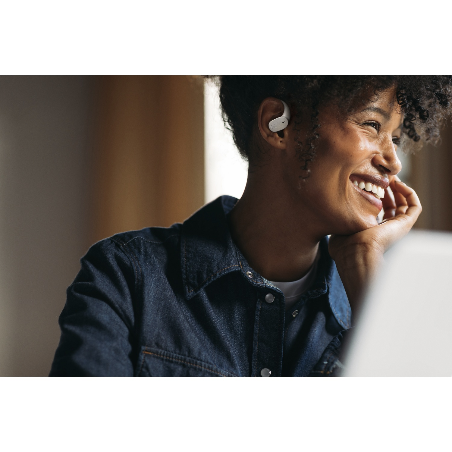 Une femme souriante avec un écouteur blanc dans l'oreille, portant une chemise en jean, posant son menton sur sa main.