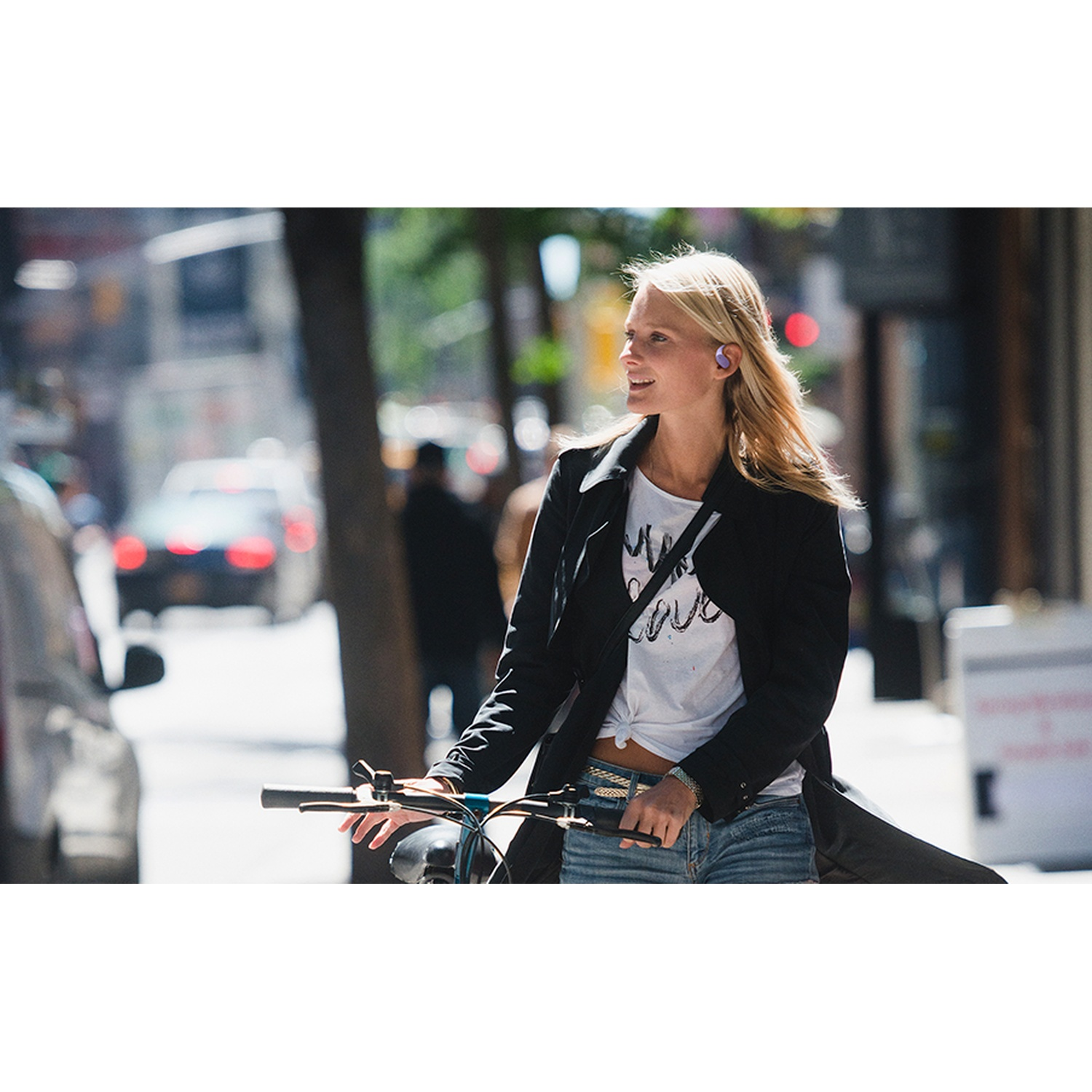 Une femme blonde porte une veste noire, une chemise blanche et un short en jean. Elle fait du vélo dans une rue de la ville avec des écouteurs.